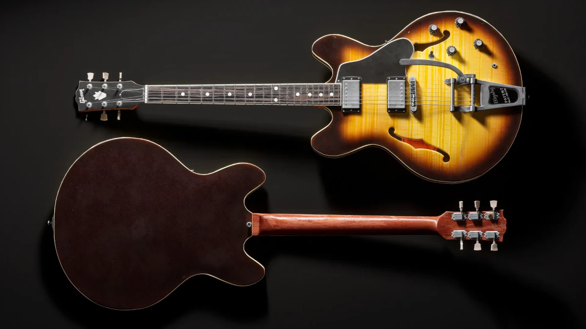 Render hiperrealista de una guitarra Gibson ES-335 en un estudio de grabación vintage. La madera muestra un desgaste natural, con reflejos cálidos en su acabado brillante. Las cuerdas ligeramente gastadas y el hardware metálico reflejan la luz ambiental. Fondos con instrumentos desenfocados y luz tenue para realismo.
